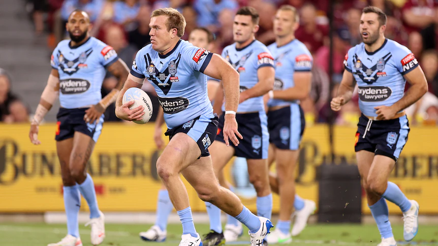 A NSW State of Origin player carries the ball as he runs forward, while his teammates stand behind him.