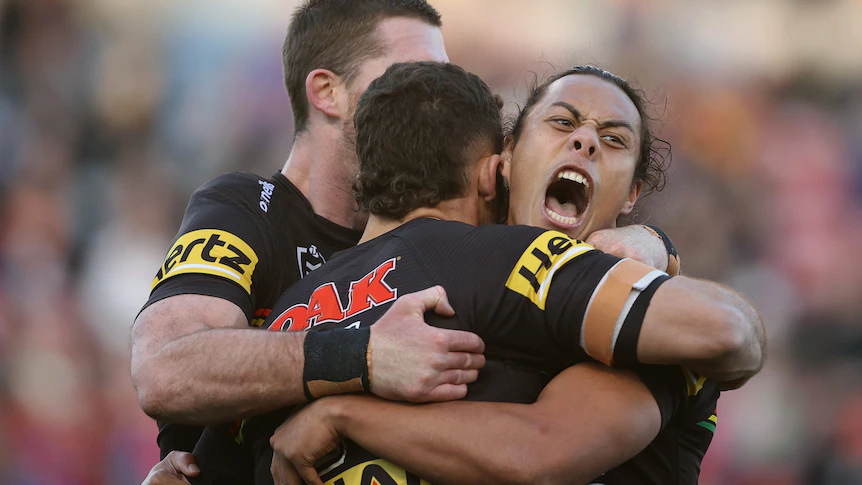 Jarome Luai shouts while being hugged by two Penrith Panthers teammates.