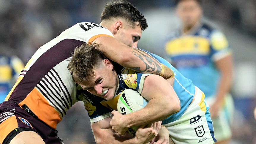 A Gold Coast Titans NRL player holds the ball as he is tackled by a Brisbane Broncos opponent.