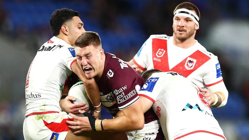 A Manly NRL player holds the ball as he is tackled by two St George Illawarra opponents.