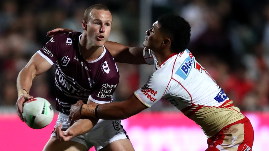 A Manly NRL player passes to his left as he is tackled by a Dolphins opponent.