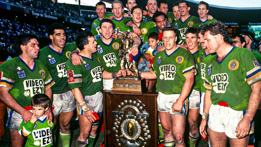 A team of men in matching green rugby jerseys stands in a group in front of a large winners plaque.