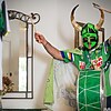A man in a green Viking face covering with horns and a drum strapped to his chest holds a drumstick in each hand above his head.