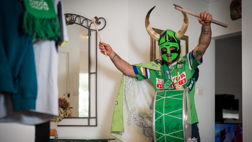 A man in a green Viking face covering with horns and a drum strapped to his chest holds a drumstick in each hand above his head.