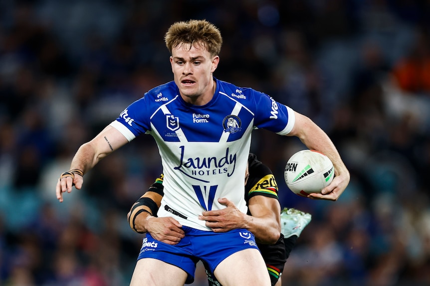 Lachlan Galvin of the Bulldogs is tackled, holding the football in one hand while standing